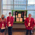 Three people in red jackets standing next to a bulletin board.