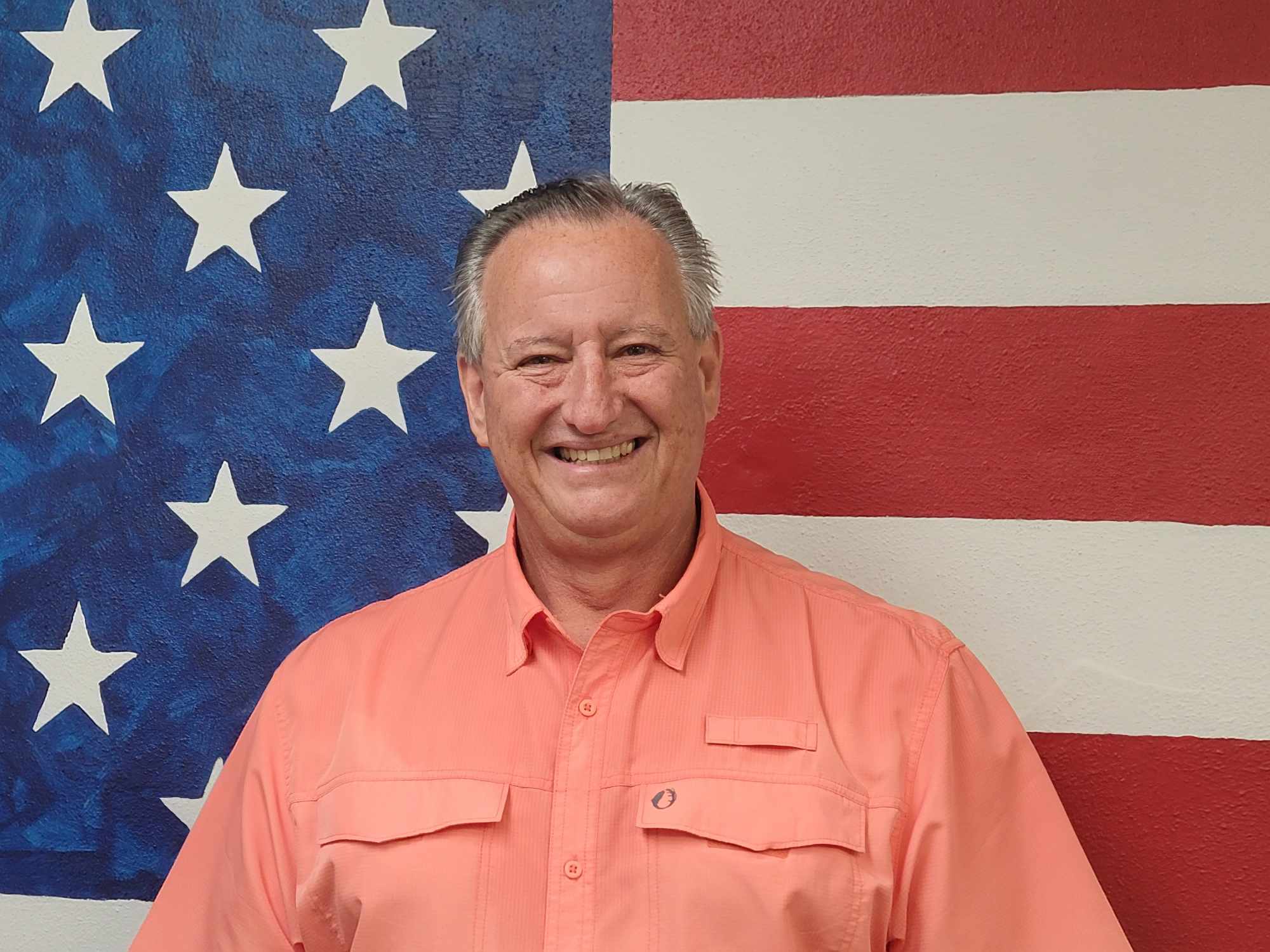 A man in an orange shirt standing next to the american flag.