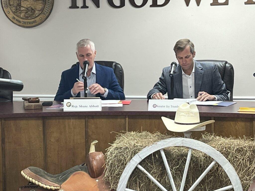 Two men sitting at a table with hay and a horse.